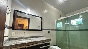 Un baño moderno con encimera de granito, un lavamanos blanco, un espejo, un inodoro y una cabina de ducha de vidrio.