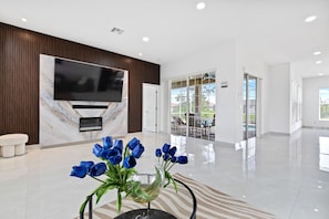 Eine moderne Lobby mit Marmorwand, einem großen Fernseher, einem Glastisch mit blauen Blumen und Blick ins Freie.