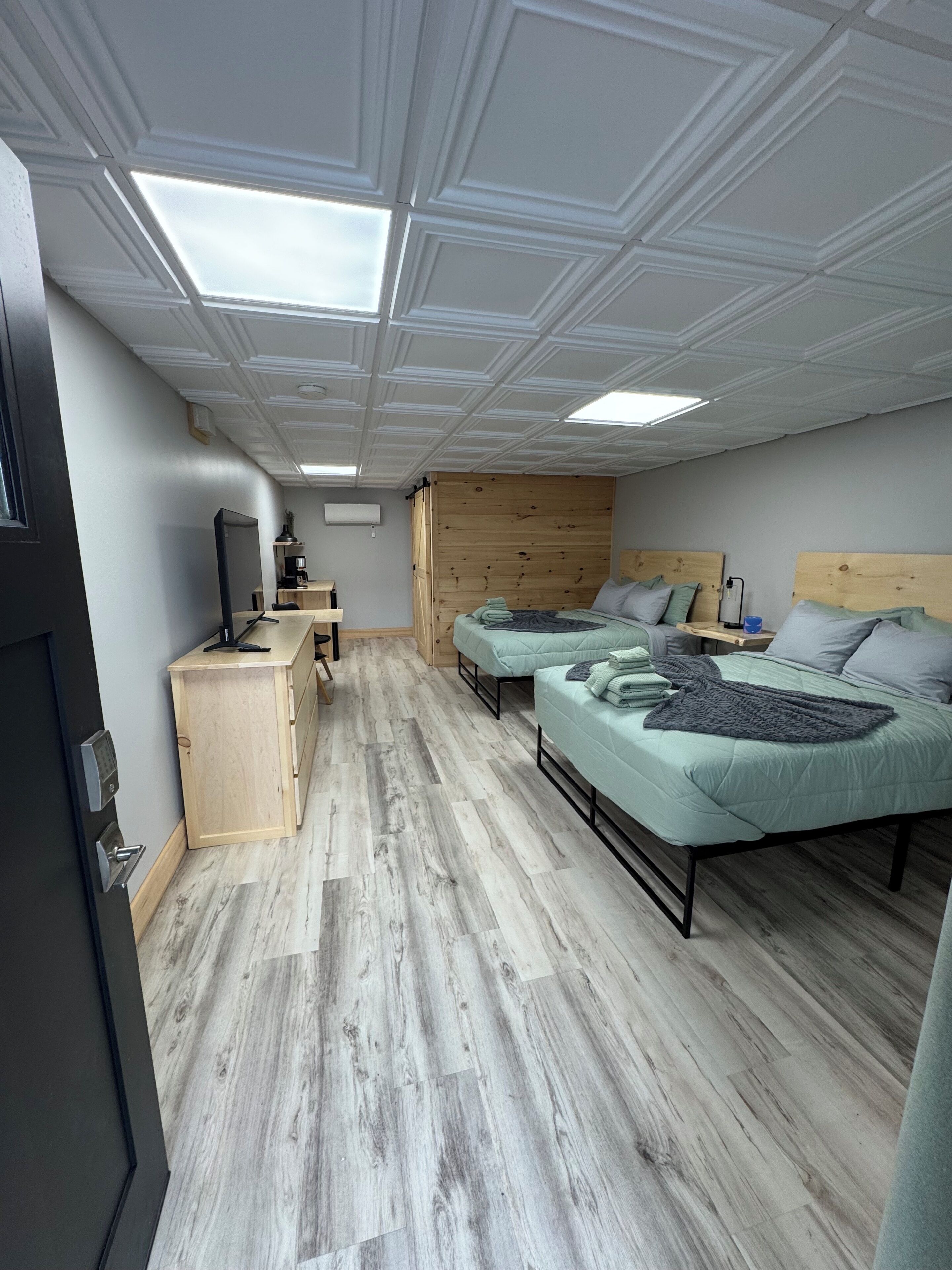 Una habitación con piso de madera, dos camas con ropa de cama verde, un escritorio con computadora y un armario de madera.