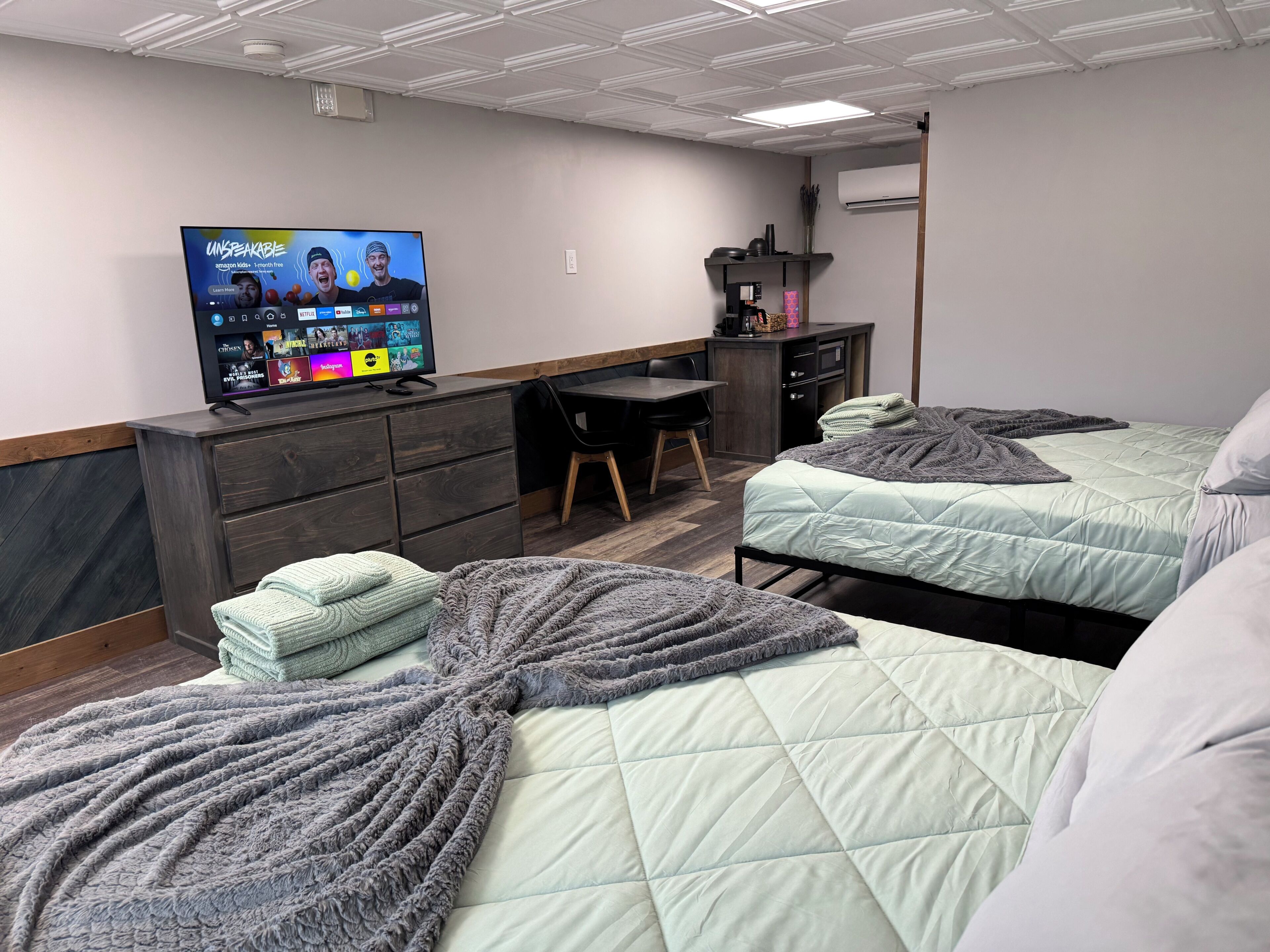 A hotel room with two beds, a desk, a TV displaying a streaming service, and a small kitchenette.