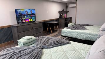 A hotel room with two beds, a desk, a TV displaying a streaming service, and a small kitchenette.