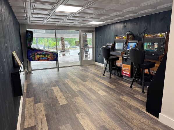 Una habitación con suelo de madera, un ventanal grande y dos máquinas de videojuegos.