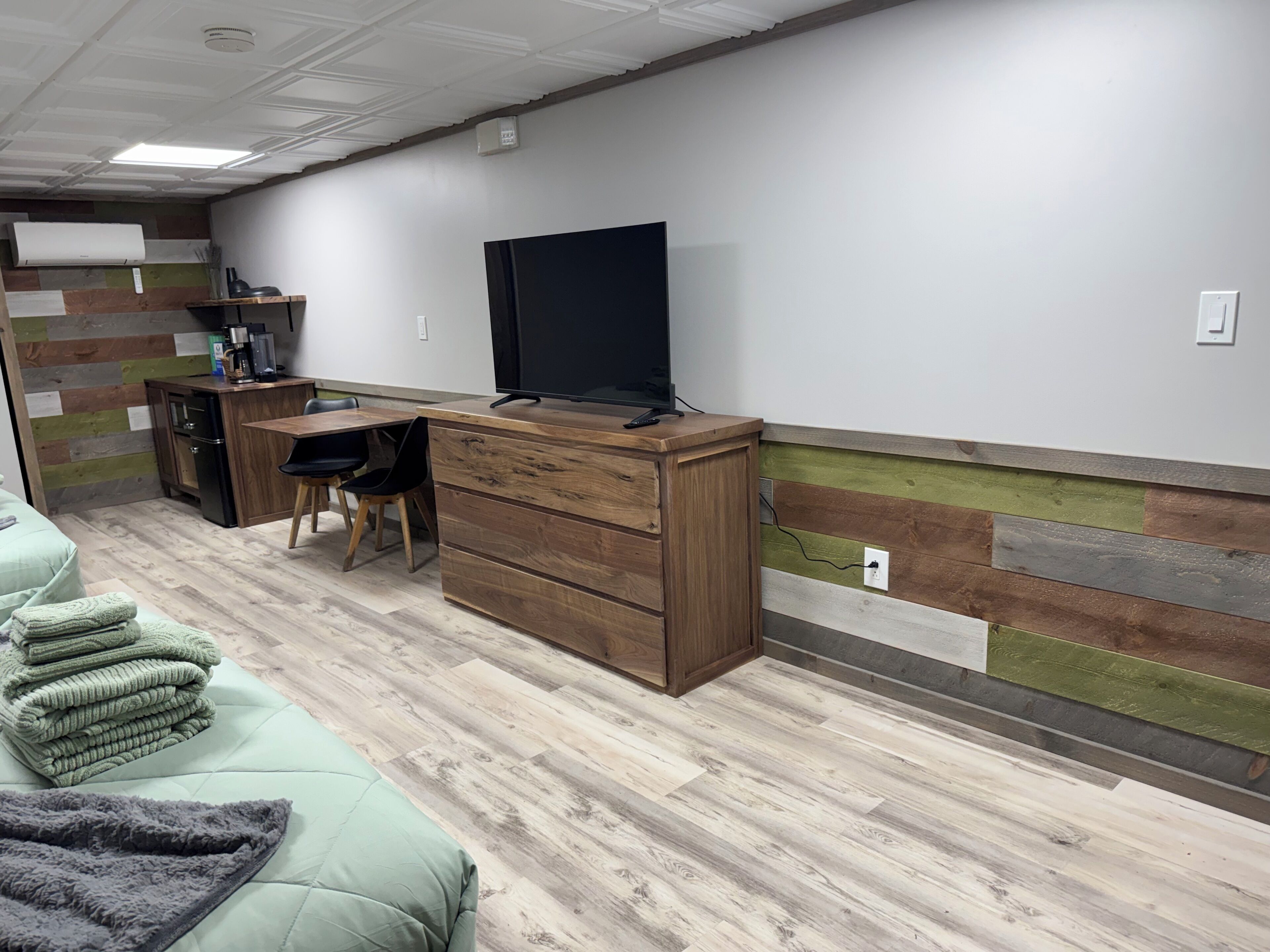 Ein Zimmer mit Holztisch, Fernseher, einer Küchenzeile und einem Bett mit grün-grauen Decken.