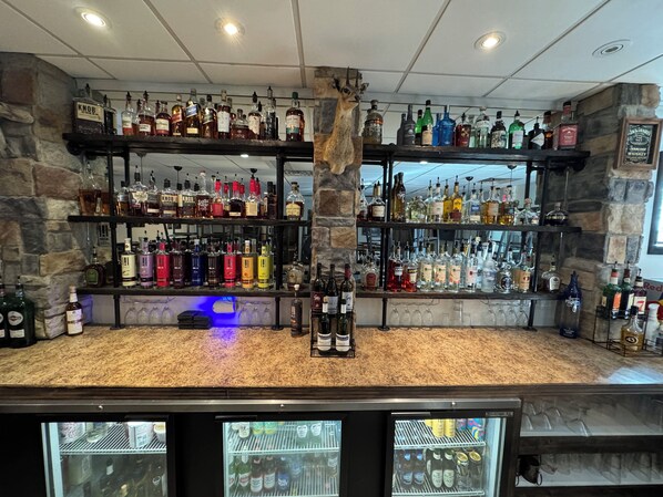Zona de bar con estantes llenos de botellas de licores, una cabeza de animal decorativa y un refrigerador con botellas.