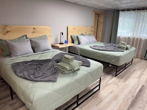 Dos camas con cabeceros, una mesita de noche con lámpara y un ventanal con cortinas.
