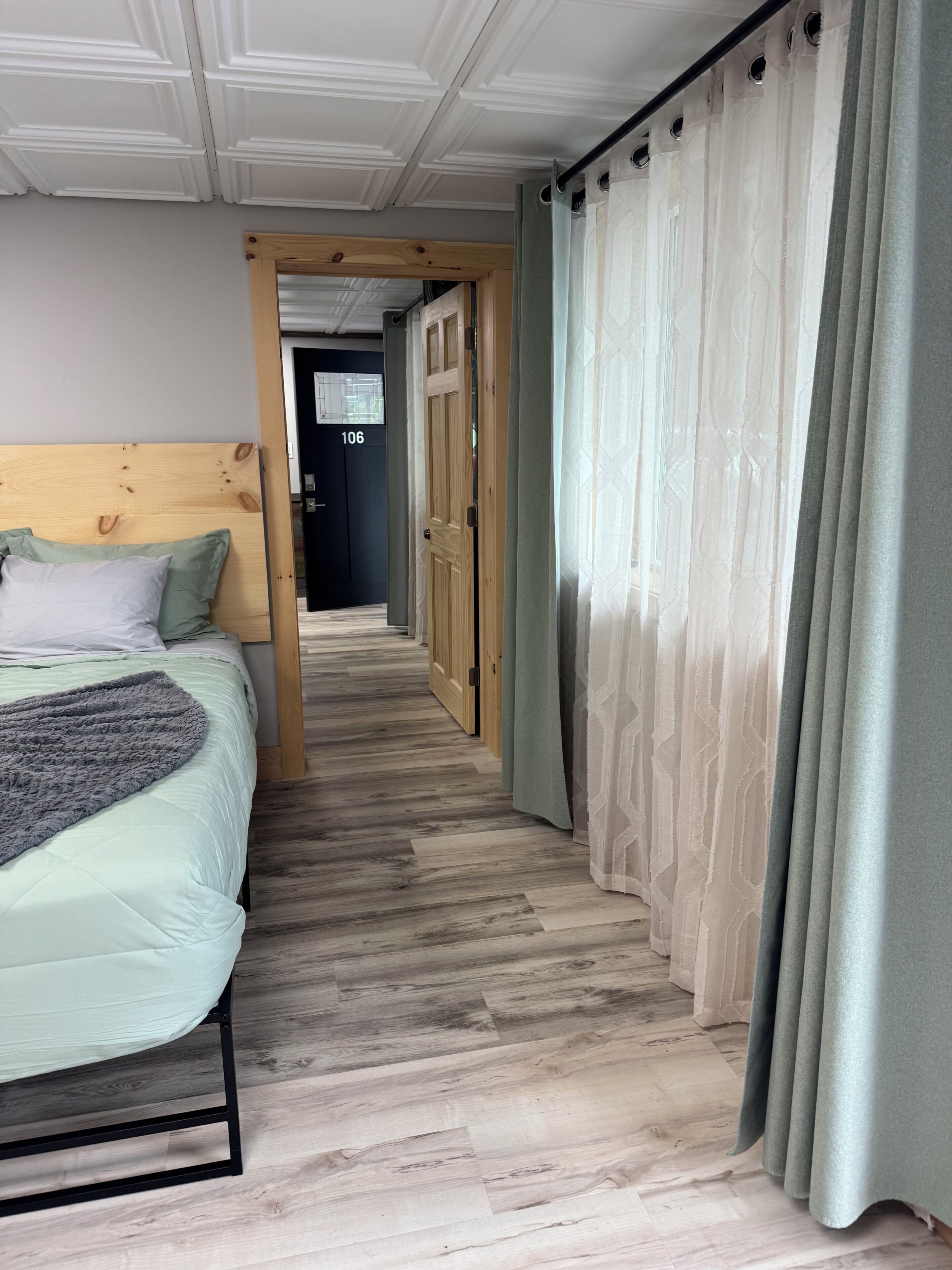 A bedroom with a bed, a wooden headboard, a grey blanket, and a hallway leading to a door marked '106'.