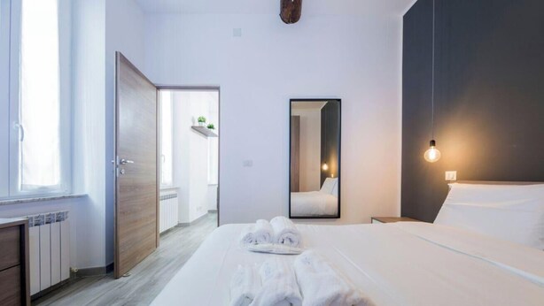 A hotel room with a bed, a bedside table, a mirror, a window, and a door leading to a bathroom. - Aquarium 3' | 2BR | A/C | Wifi & Netflix by Tilo (Genoa)