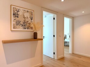A hallway with a framed floral artwork, a wooden shelf with a vase, and a door leading to another room. - Barreiro Nooba 263C Apartment BY LULUNA (Barreiro)