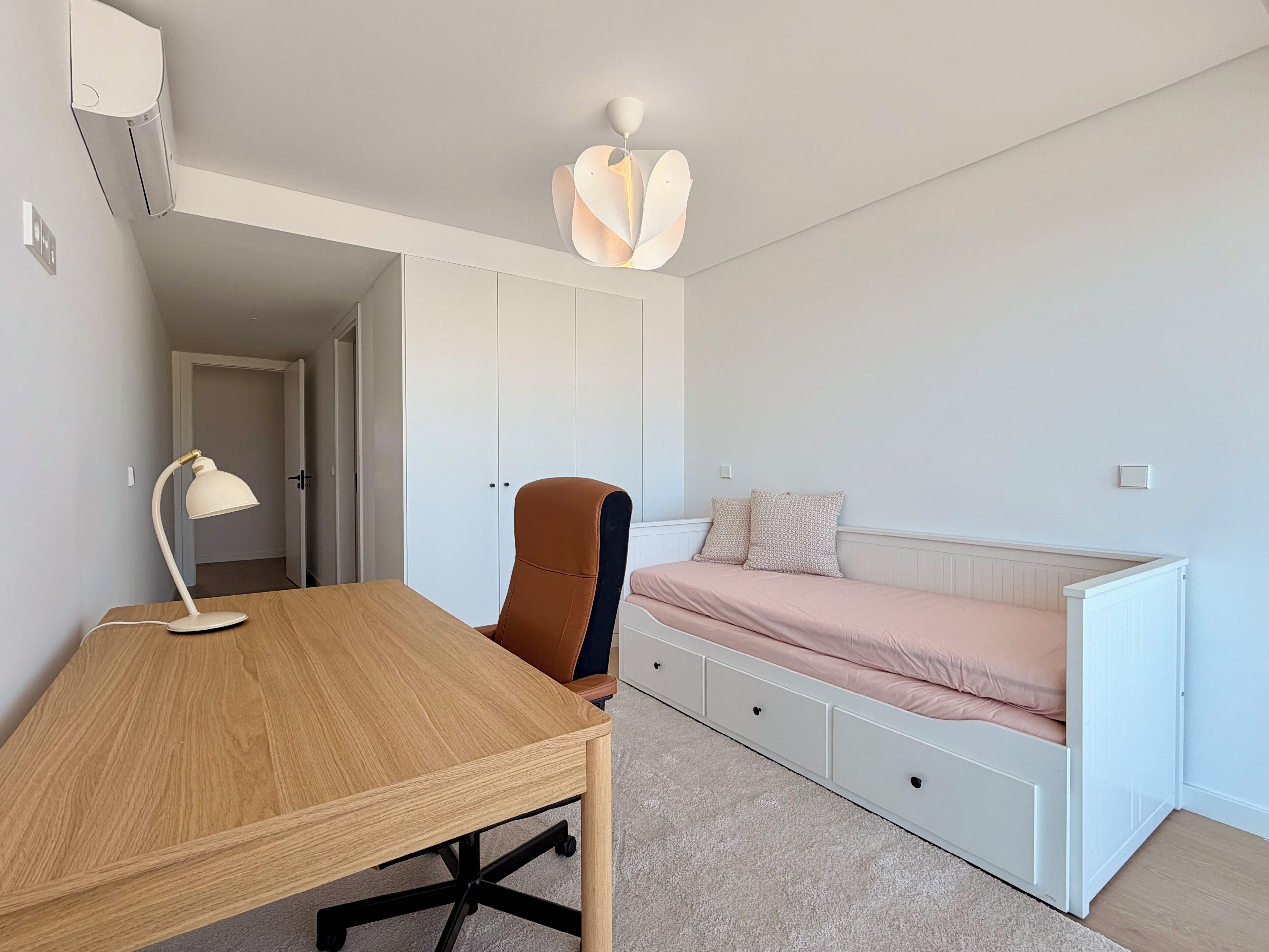 Una camera da letto con una scrivania in legno, una sedia, un letto con materasso rosa e un armadio bianco, oltre a una lampada a soffitto.