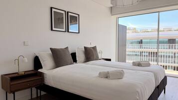 A modern hotel room with a large bed, two bedside tables, a nightstand, a large window with a view, and two framed pictures on the wall.
