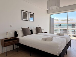 Une chambre d’hôtel moderne avec un grand lit, deux tables de chevet, une petite table d’appoint, une grande fenêtre donnant sur un beau paysage et deux tableaux encadrés accrochés au mur.