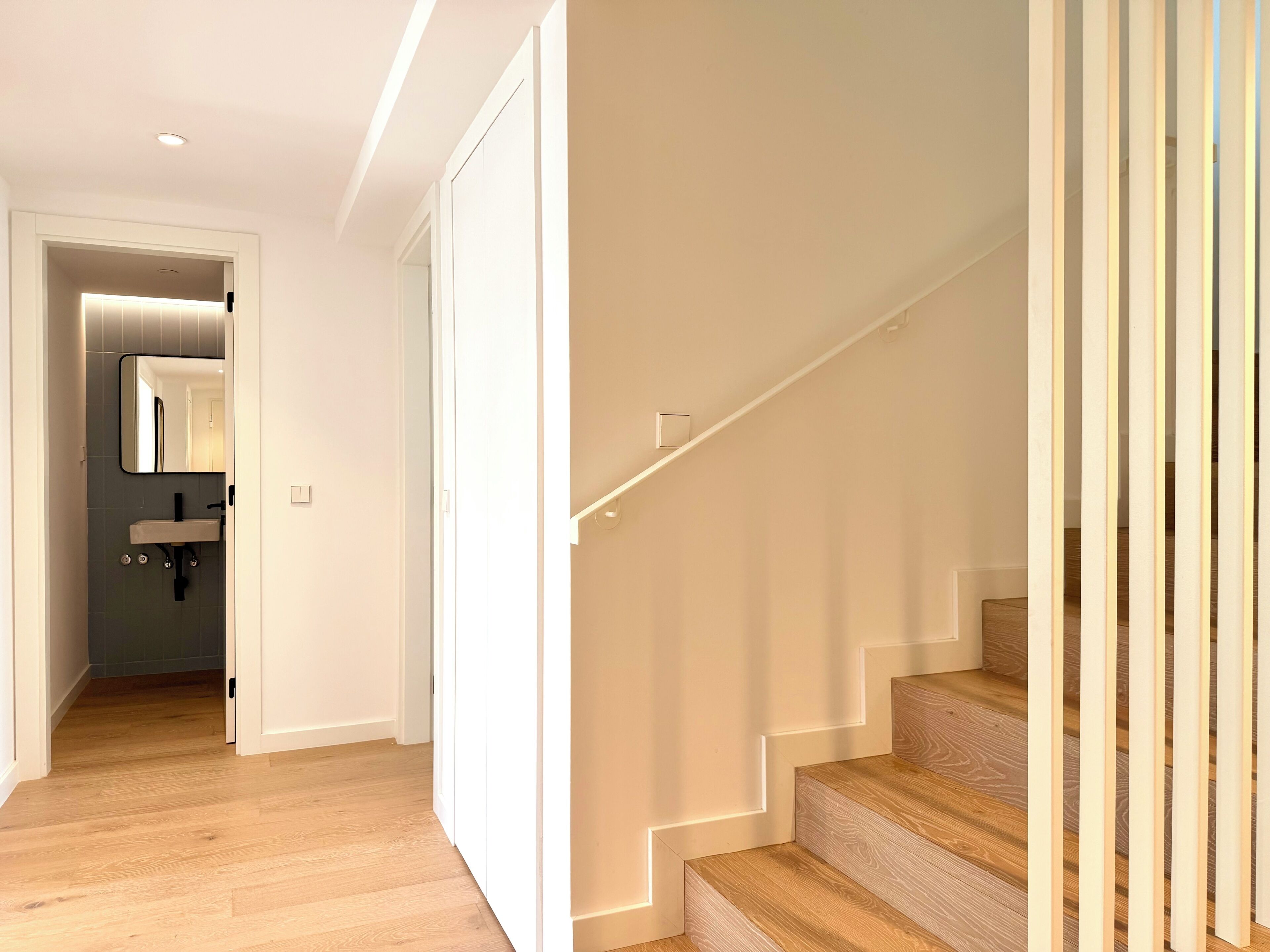 A modern interior with a staircase, a hallway leading to a bathroom, and a light-colored floor.