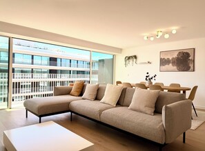A modern living room with a sectional sofa, a dining table with chairs, a large window with a view of a building, and a ceiling light fixture. - Barreiro Nooba 263C Apartment BY LULUNA (Barreiro)