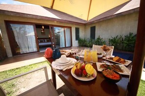 A breakfast table with plates of food, drinks, and a view of a patio with a sliding door. - Pousada Rangai (Maragogi)