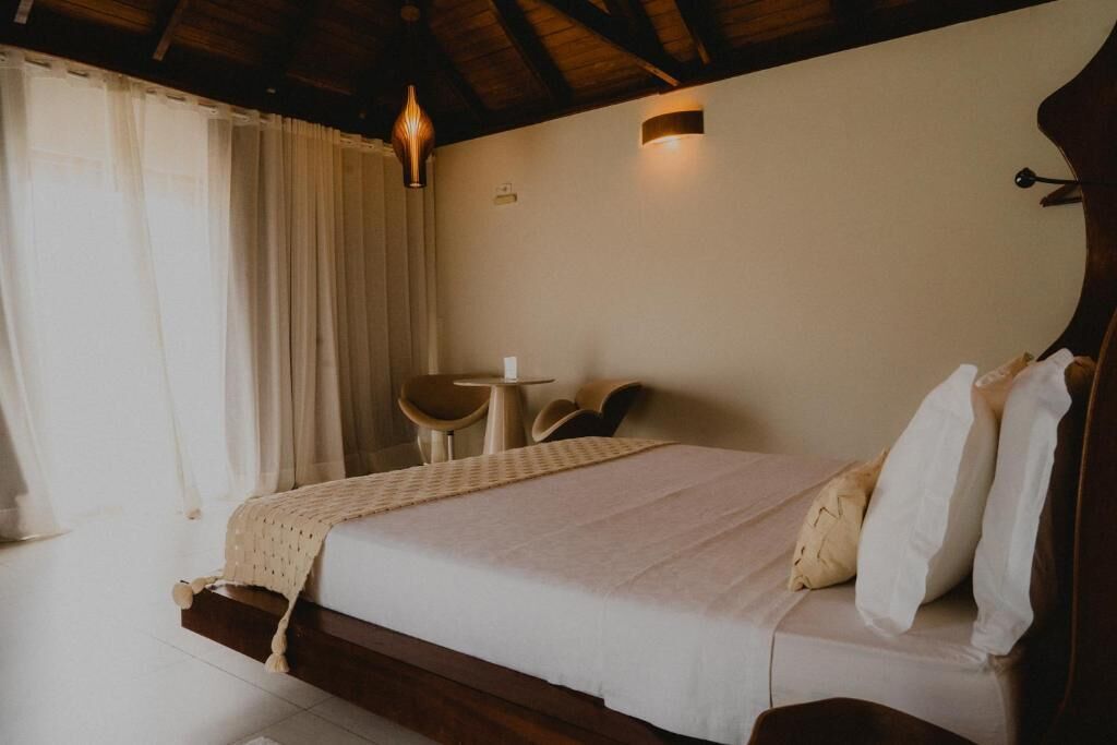 A bedroom with a bed, a small table, two chairs, a lamp, and a window with curtains.
