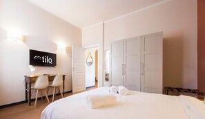 A hotel room with a bed, a desk with chairs, a wardrobe, and a wall-mounted TV. - Aquarium 3'|2BR|Lift|Wifi|Train Station|By Tilo (Genova)