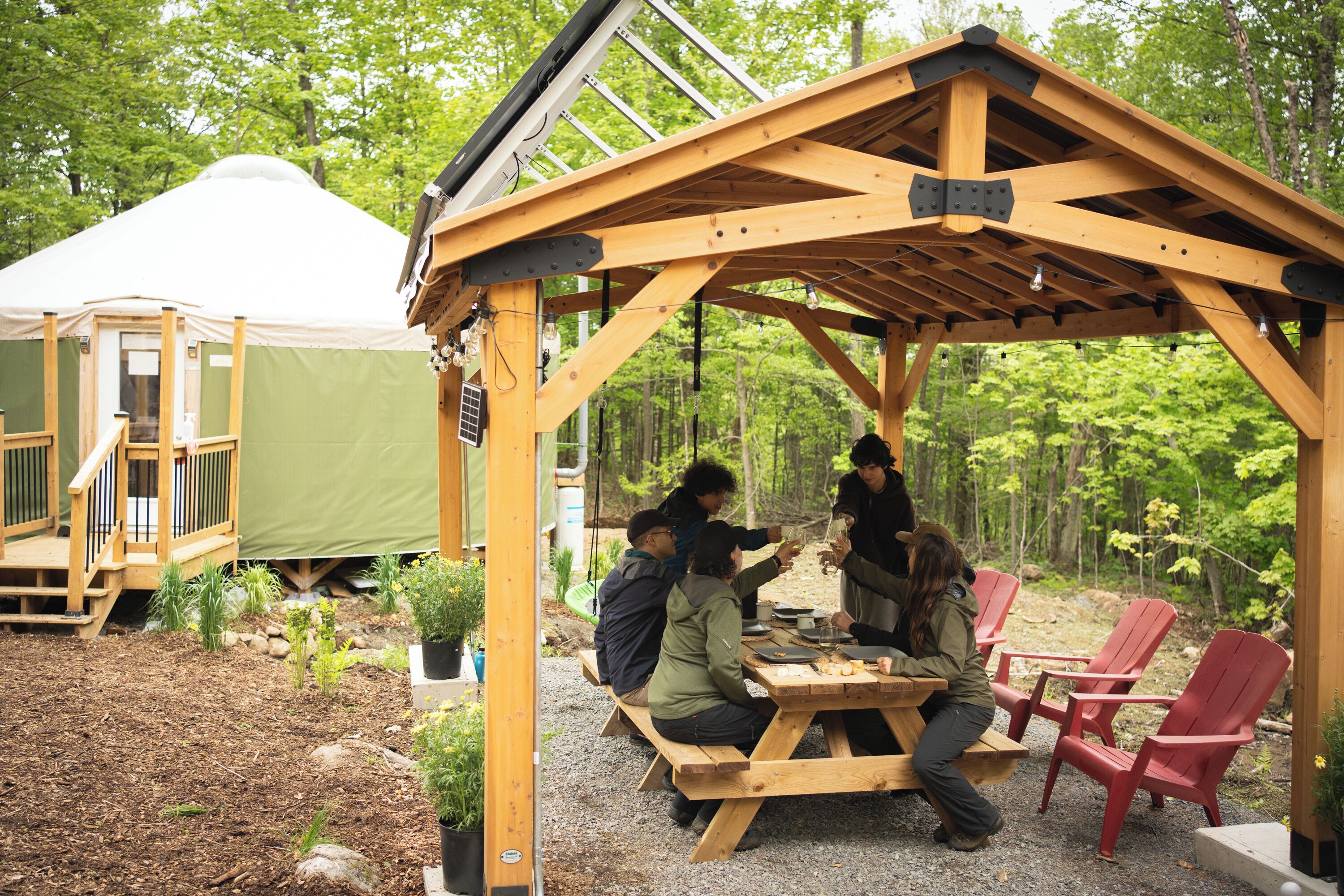 Un kiosque en bois avec une table de pique-nique et des chaises rouges, entouré de verdure et d’une tente.