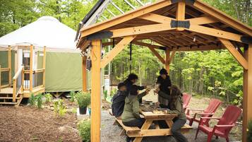 Un kiosque en bois avec une table de pique-nique et des chaises rouges, entouré de verdure et d’une tente.