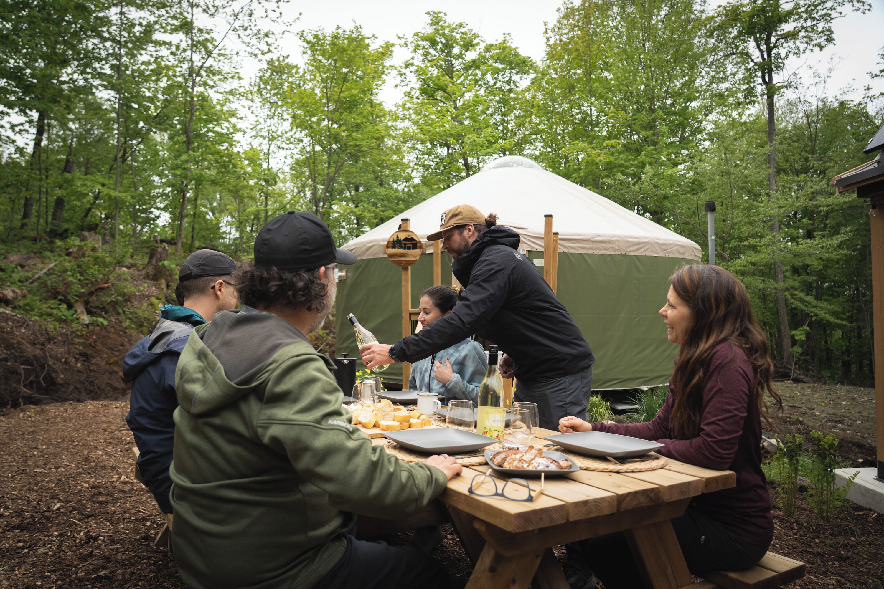 Eine Gruppe von Personen sitzt an einem Holztisch im Freien, im Hintergrund ist ein Zelt zu sehen.