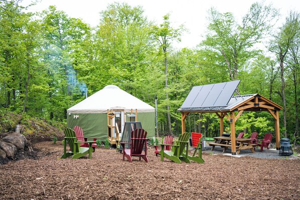 Ein Waldcampingplatz mit Zelt, einer überdachten Sitzgruppe und mehreren Adirondack-Stühlen.
