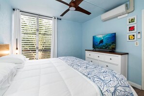A bedroom with a bed, a ceiling fan, an air conditioning unit, a television, and a window with blinds. - Alii Villas#221 (Kailua-Kona)