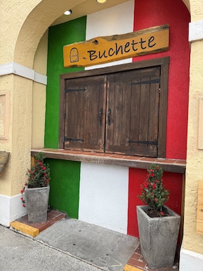 Eine Gebäudefassade mit einem Schild mit der Aufschrift 'Buchette', einer Holztür und zwei mit roten Blumen bepflanzten Blumentöpfen.
