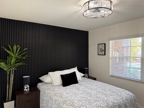 Ein Schlafzimmer mit Bett, einer Topfpflanze, einer Lampe, einem Bilderrahmen und einem Fenster mit Jalousien.