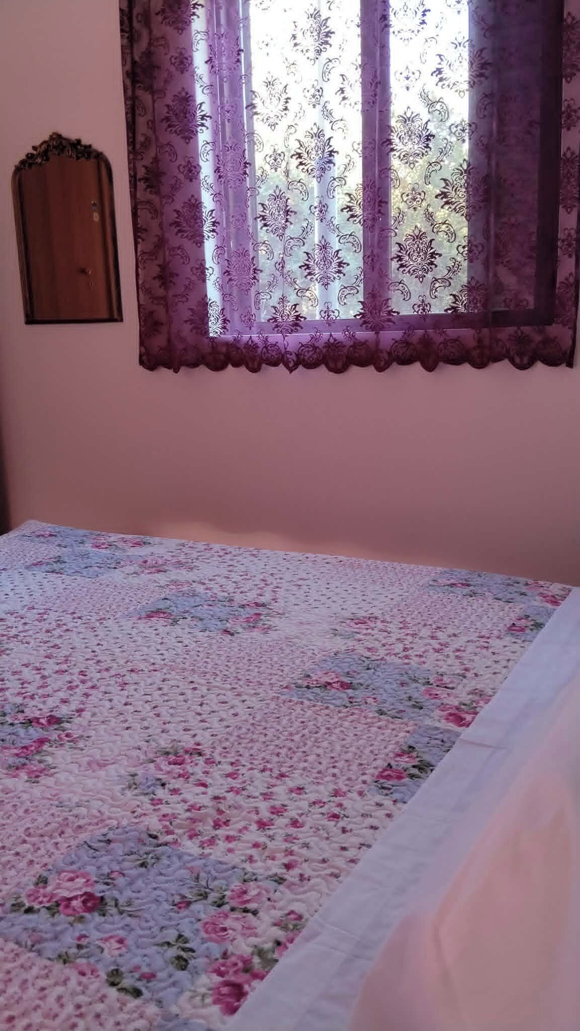A bedroom with a floral bedspread, a window with purple curtains, and a wooden door.