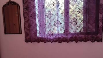A bedroom with a floral bedspread, a window with purple curtains, and a wooden door.