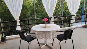 Ein runder Tisch mit weißen Spitzen, zwei schwarze Stühle, eine Vase mit einer rosa Blume und weiße Vorhänge an einem Balkon mit Geländer.