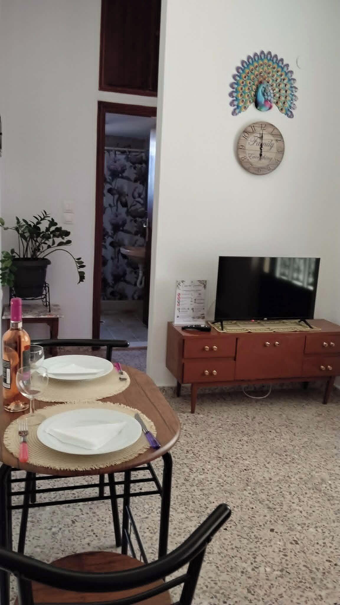 Ein Essbereich mit Tisch für zwei Personen, einem Fernseher auf einem Holzschrank und einer dekorativen Wanduhr.