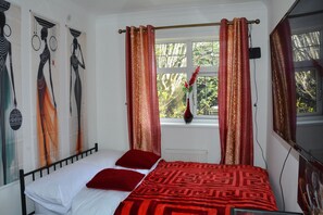 Une chambre à coucher avec un lit, une literie rouge et blanche, une fenêtre avec des rideaux, un vase de fleurs et des œuvres d’art mural.
