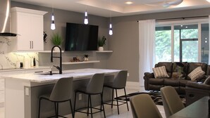 A modern kitchen with a central island, bar stools, a flat-screen TV, and a dining area with a brown leather couch. - Family Friendly SWFL Home• 3BR, Sleeps 6, Screened Lanai (Fort Myers)