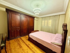 Ein Schlafzimmer mit Holzboden, einem Bett mit rosa Bettdecke, einem Holzschrank, einem Stuhl und einer Deckenleuchte.