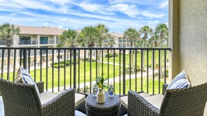 A balcony with wicker furniture, a small table, and a view of a residential area with palm trees and a building. - Ocean Views, Two Balconies, Easy Beach Access, Two Pools, 5 miles to Downtown, $300 in Free Tickets! (St. Augustine Beach)