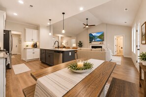 Et moderne køkken med en central ø, et spiseområde med et træbord, og en stue med sofa og pejs.