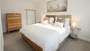 A bedroom with a large bed, a wooden dresser, a bedside table with a lamp, and a wall-mounted abstract painting. - Modern 3BR Home - Super King, Office & Garden (Hemel Hempstead)