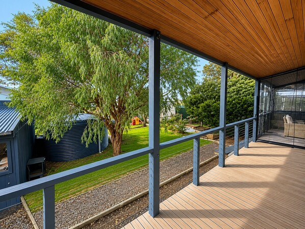A covered outdoor area with a wooden deck, metal railings, and a view of a garden with trees and a small building.