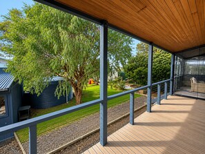 A covered outdoor area with a wooden deck, metal railings, and a view of a garden with trees and a small building.