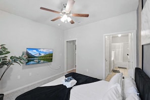 Un dormitorio con cama, ventilador de techo, televisor en la pared y puerta que da a un baño.