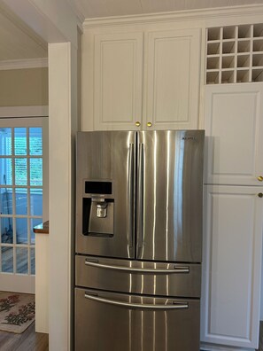 Un refrigerador d'acer inoxidable amb dispensador d'aigua, ubicat a la cuina que té armaris blancs i una finestra amb persianes.