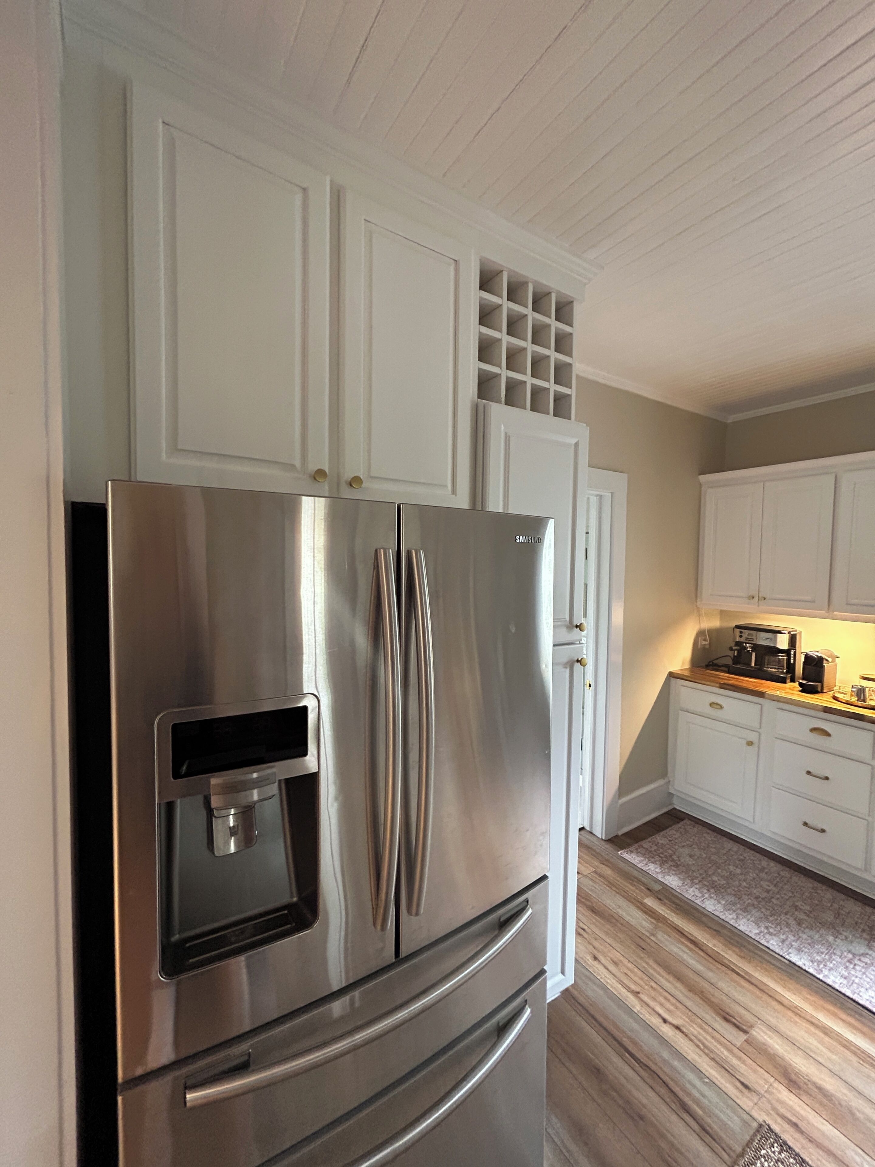 A stainless steel refrigerator with a water dispenser, white cabinetry, and a wooden floor in a kitchen.