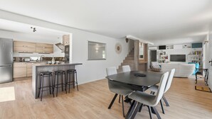 A modern kitchen with a central island, a dining area with a dark table and chairs, and a living area with a sofa and bookshelves.