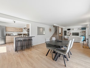 Une cuisine moderne dotée d’un îlot central, un coin repas avec une table et des chaises aux couleurs sombres, et un espace de vie comprenant un canapé et des étagères.