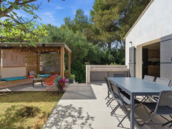 A patio area with a dining table and chairs, a wooden deck with a seating area, and a kitchen visible through an open door.