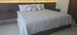Ein Bett mit weißer Bettdecke, dekorativen Kissen, einem Holz-Kopfende und einer Wand aus Stein mit warmem Licht.