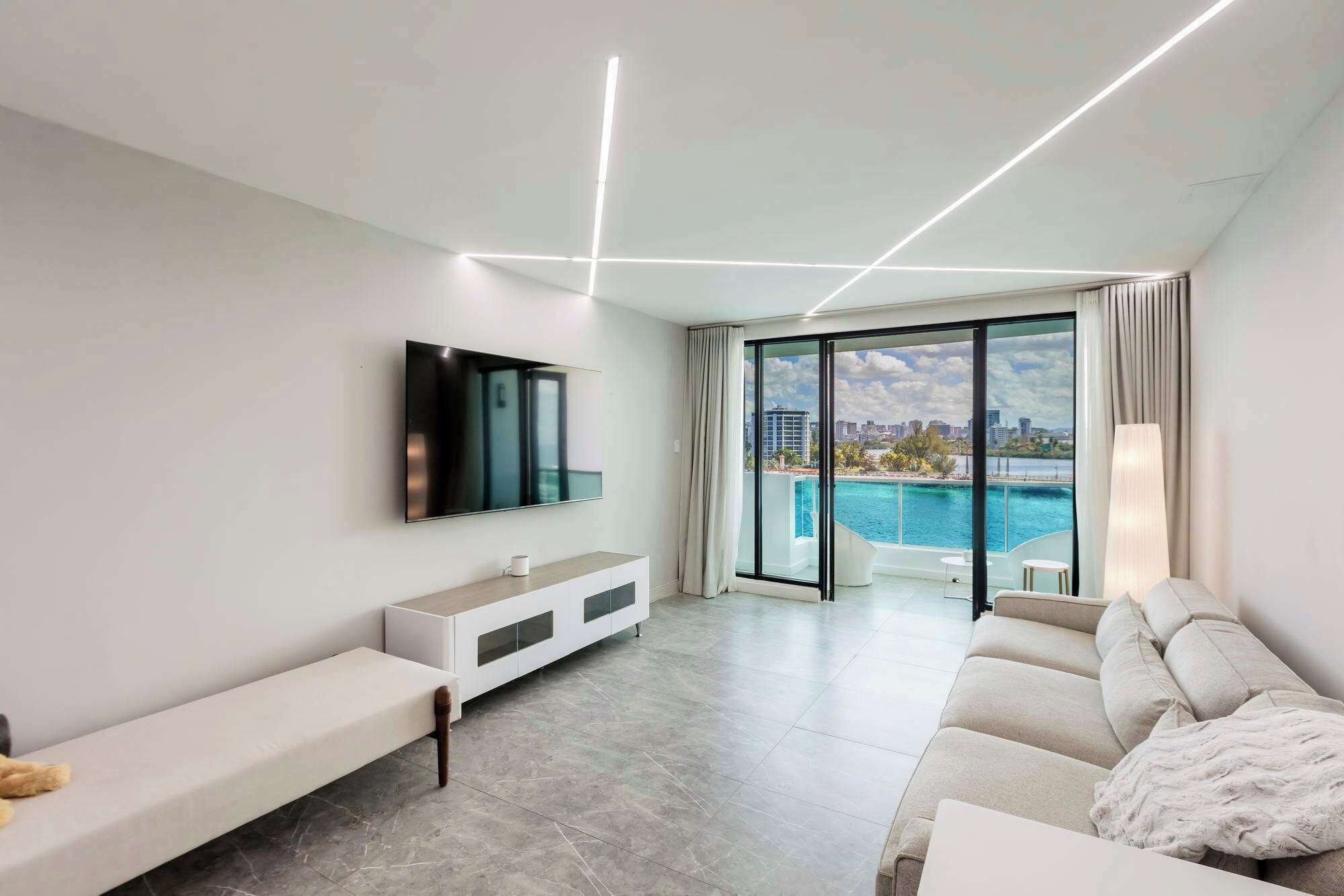 A modern living room with a flat-screen TV, a sofa, a coffee table, and a view of a swimming pool.