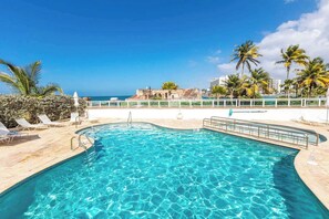 Una piscina con agua azul cristalina, rodeada de sillas de descanso y palmeras, con vista al océano y edificios al fondo.