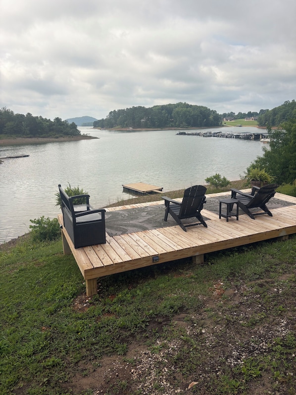 Una terraza de madera con mobiliario exterior con vista a un lago tranquilo, rodeada de vegetación y colinas al distante.