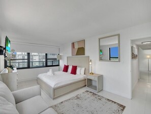 A modern hotel room with a large bed, a bedside table, a mirror, and a view of the city through the window. - Spacious Corner 2 BDR Condo Beachfront Resort Beach Service, Pools, Gym 1020 (Miami Beach)